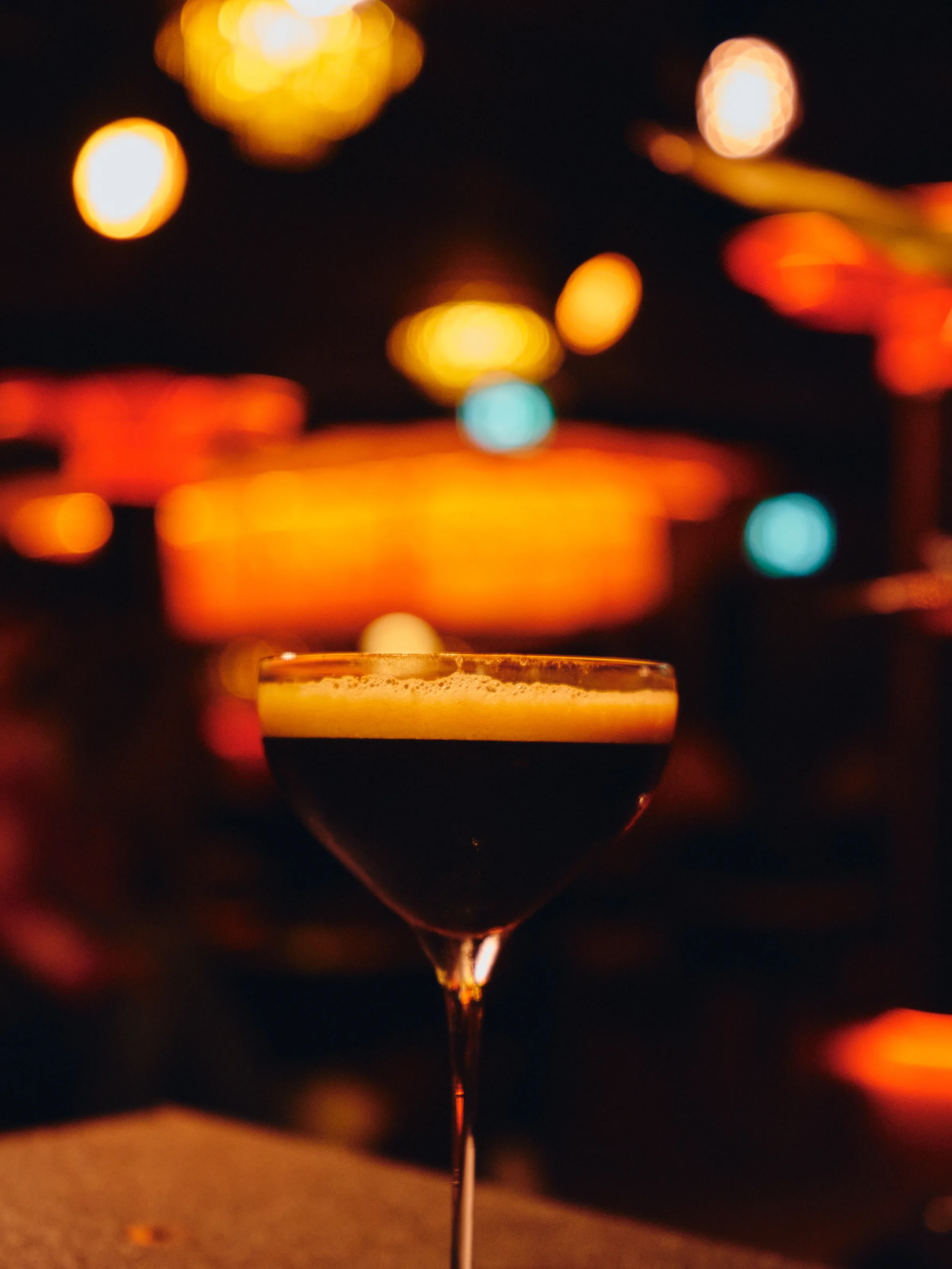 Mørk cocktail med kremet skum i martiniglass mot varmt bokeh-lys